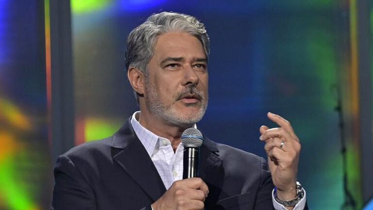 Jornalista William Bonner de terno escuro e camisa clara segura microfone prata enquanto gesticula com a mão esquerda em estúdio com luzes coloridas