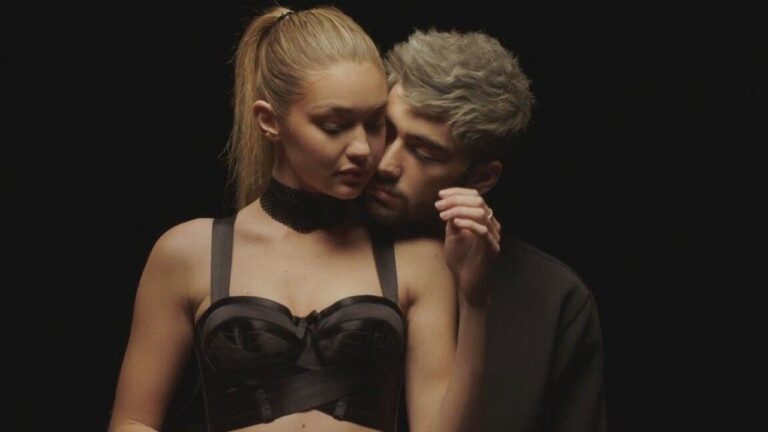 Casal posa em clima intimista contra fundo escuro, com o homem abraçando a mulher por trás enquanto se aproximam em gesto de carinho; ambos vestem roupas pretas sob iluminação suave e dramática