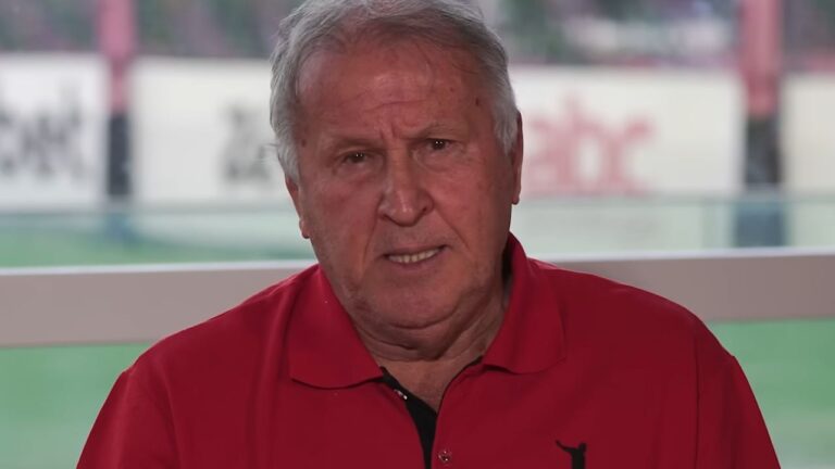Ex-jogador Zico veste camisa polo vermelha e olha para a câmera com expressão séria, sentado em um ambiente com fundo desfocado que mostra um campo