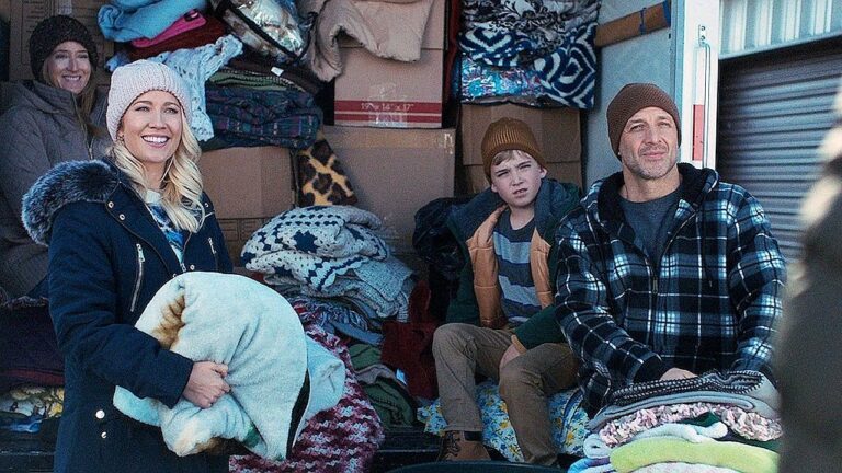 Quatro pessoas estão sentadas e em pé na parte traseira aberta de um caminhão ou veículo de carga cheio de caixas e cobertores empilhados. Elas vestem roupas de frio, incluindo casacos, gorros e luvas. Uma mulher segura um travesseiro enquanto sorri, um homem e um garoto estão sentados ao lado de pilhas de cobertores dobrados, e outra mulher aparece ao fundo. A cena sugere um ambiente frio e possivelmente uma atividade de transporte ou distribuição de roupas e mantas.