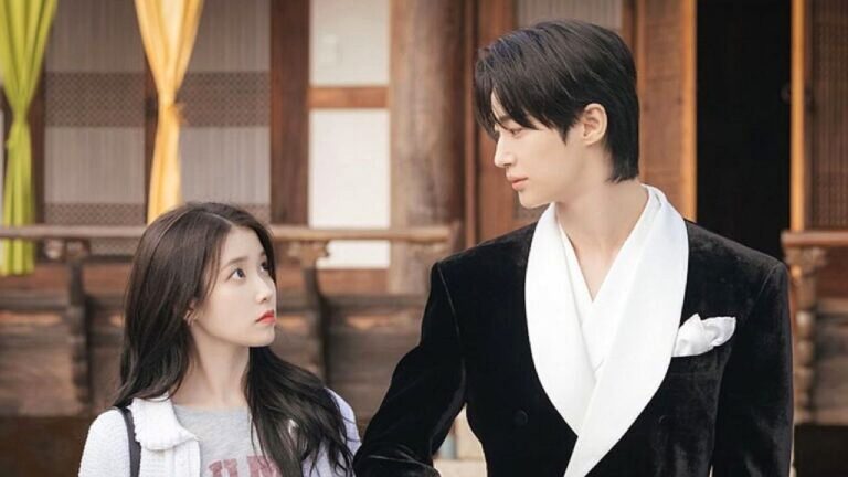 Um homem alto de cabelo escuro, vestindo terno preto com camisa branca, caminha ao lado de uma mulher jovem de cabelos longos; ela olha para ele com expressão curiosa enquanto estão diante de uma construção de madeira ao fundo.