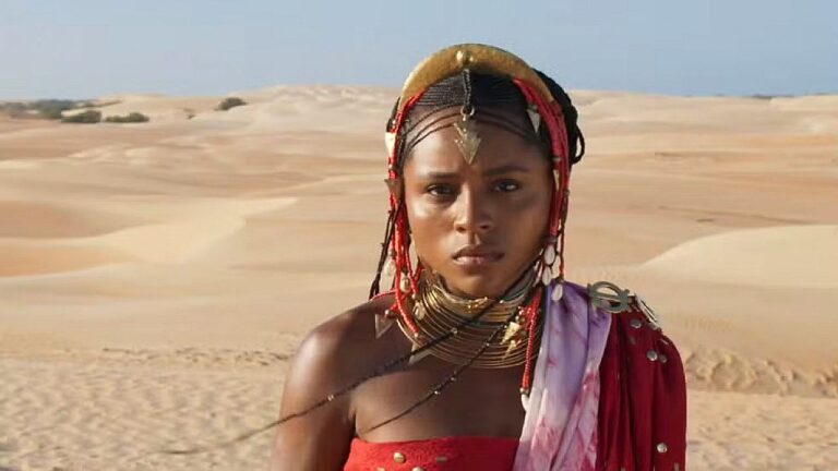 Mulher em um cenário de deserto, usando roupas e adornos tradicionais com colares e enfeites na cabeça, olhando seriamente para a frente.