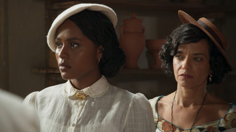 Duas mulheres com roupas de época e chapéus observam algo com expressão séria em cena da novela A Nobreza do Amor.
