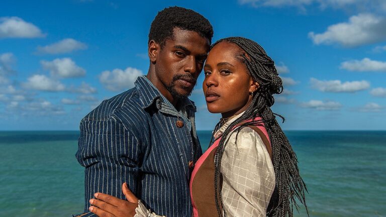 Um homem e uma mulher estão próximos um do outro, posando diante do mar. O homem tem cabelo curto e barba e veste uma camisa azul com listras. A mulher tem cabelo trançado e usa roupas em tons neutros com detalhes rosados. Ambos olham diretamente para a câmera com expressões sérias. Ao fundo, aparece o oceano e um céu azul com algumas nuvens, sugerindo um ambiente costeiro ao ar livre.