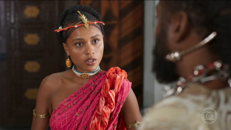Mulher com tranças e adorno dourado na cabeça usa vestido rosa com detalhes e joias enquanto conversa com outra pessoa em cena de A Nobreza do Amor; ao fundo aparece o logotipo da Globo.