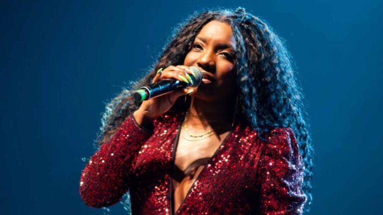 Adriana Araújo se apresenta no palco segurando um microfone, vestindo blazer vermelho com brilho e decote profundo. Ela tem cabelos longos e volumosos e está iluminada por luz azul de fundo durante o show