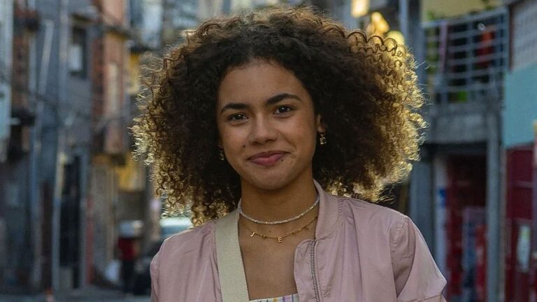 Mulher jovem de cabelos cacheados volumosos sorri levemente enquanto caminha por rua urbana, usando jaqueta rosa e colares delicados, com prédios e casas desfocados ao fundo.