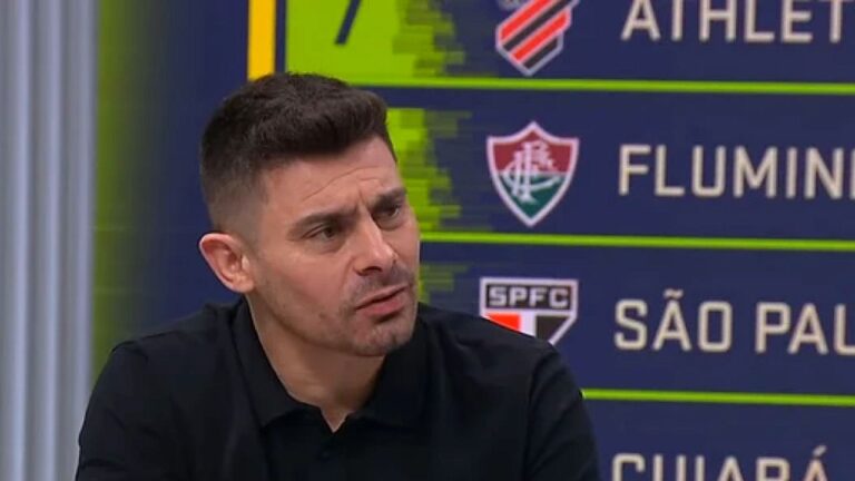 Homem de cabelo curto e barba por fazer participa de programa esportivo em estúdio de TV, olhando para o lado enquanto fala; ao fundo, um painel exibe nomes e escudos de clubes de futebol brasileiros, como Fluminense e São Paulo.