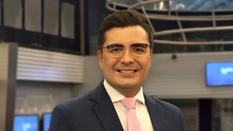 Um homem aparece em um ambiente interno moderno, sorrindo diretamente para a câmera. Ele usa óculos, terno escuro, camisa branca e gravata rosa clara, com aparência formal. O fundo sugere um estúdio ou espaço corporativo, com iluminação suave, estruturas metálicas e telas desfocadas. A composição transmite um contexto profissional, possivelmente ligado à televisão ou ao setor empresarial.