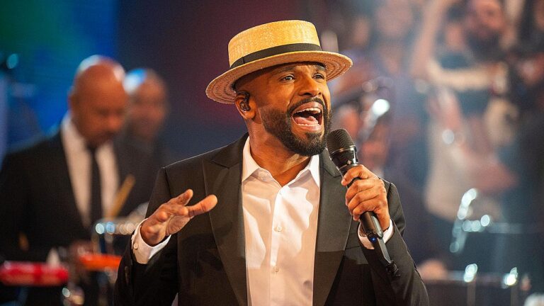 Homem cantando no palco e segurando um microfone, usando terno escuro e chapéu claro, com músicos ao fundo e iluminação de show.