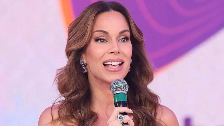 Mulher sorri e fala ao microfone durante apresentação em programa de TV com cenário colorido ao fundo