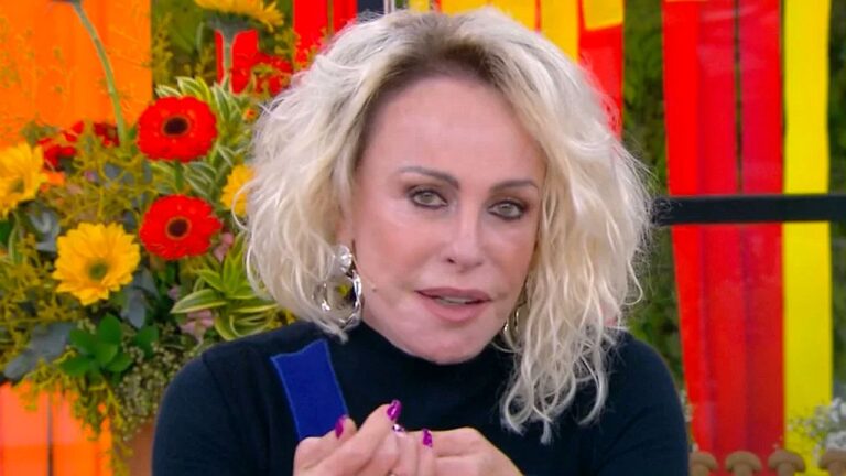 Uma mulher de cabelos loiros volumosos aparece em primeiro plano em um estúdio de televisão. Ela usa uma blusa preta de gola alta e brincos grandes e chamativos. Sua maquiagem é marcante e suas unhas estão pintadas de roxo. Ao fundo, há um arranjo de flores coloridas, incluindo flores vermelhas e amarelas, e painéis decorativos em tons de vermelho, amarelo e laranja, compondo um cenário vibrante típico de programa de TV. Ela parece estar falando enquanto gesticula com as mãos.