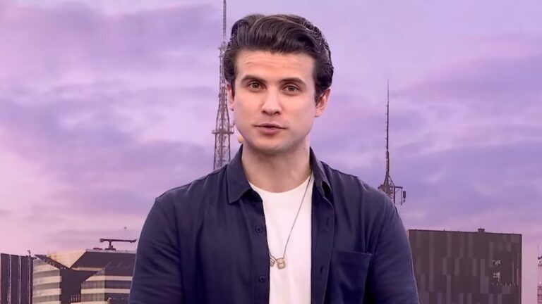 Apresentador André Marinho veste camisa azul marinho sobre camiseta branca, usa colar e posa em frente a torres de transmissão sob um céu roxo de fim de tarde