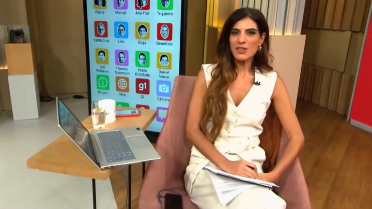 Andréia Sadi, apresentadora do GloboNews, sentada com laptop e telão do G1 ao fundo durante o Estúdio i.