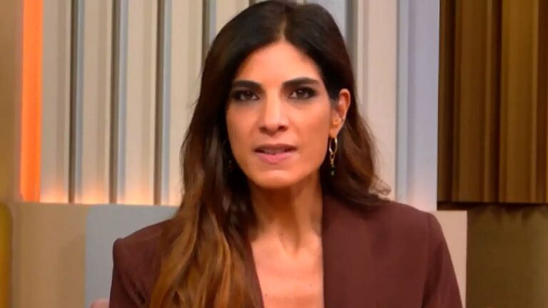 Apresentadora de telejornal usa blazer marrom e fala ao vivo em estúdio com painel iluminado ao fundo