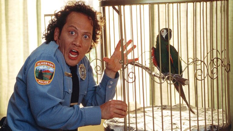 Um homem usando uniforme de policial aparece ao lado de uma gaiola com um papagaio. Ele tem expressão exagerada de surpresa, com olhos arregalados e boca aberta, enquanto levanta a mão próxima à gaiola. O pássaro está empoleirado dentro da estrutura metálica. O ambiente parece interno e bem iluminado, sugerindo uma cena de tom cômico.