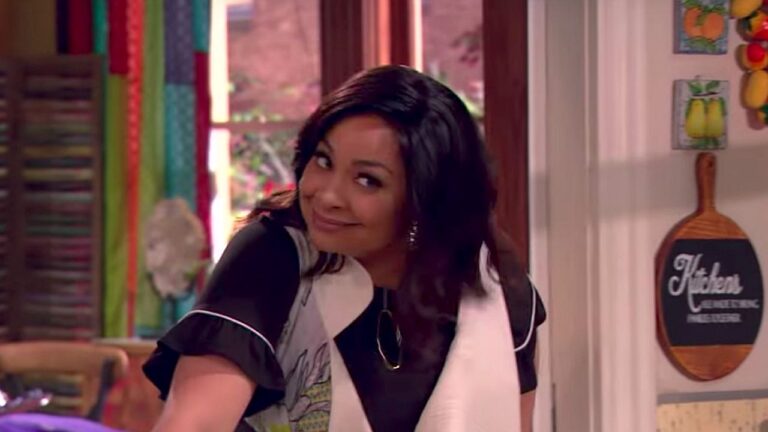 Uma mulher jovem de cabelo escuro e comprido está inclinada levemente para frente e sorrindo com expressão divertida. Ela usa uma blusa preta com detalhes claros e brincos pequenos. A cena acontece em uma cozinha colorida, com janela ao fundo, cortinas vibrantes e utensílios ou placas decorativas na parede, incluindo uma placa com a palavra “Kitchen”.