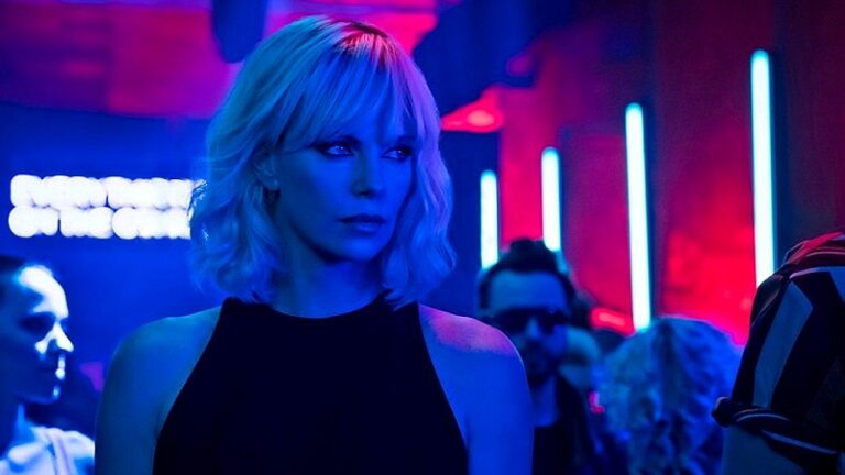 Uma mulher de cabelo curto e loiro aparece em um ambiente noturno com iluminação intensa em tons de azul e vermelho, semelhante a uma boate ou bar. Ela veste uma blusa preta sem mangas e olha para o lado com expressão séria e atenta. Ao fundo, há outras pessoas desfocadas e luzes verticais de neon, reforçando o clima urbano e noturno da cena.