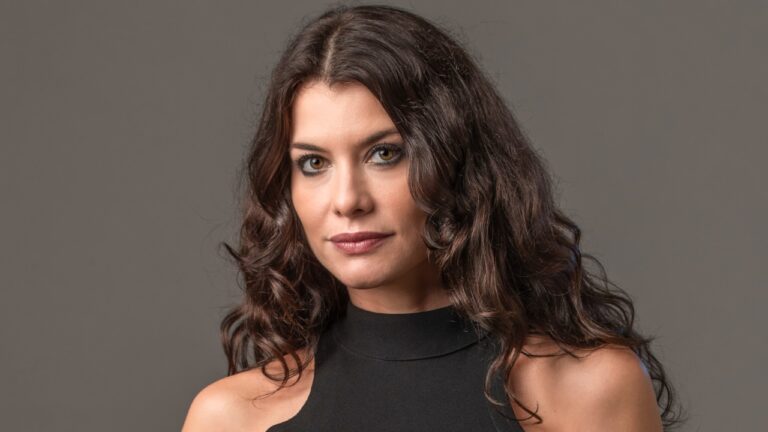Alinne Moraes interpreta Bárbara na novela Um Lugar ao Sol, exibe longos cabelos escuros e ondulados, veste blusa preta de gola alta, fundo cinza claro