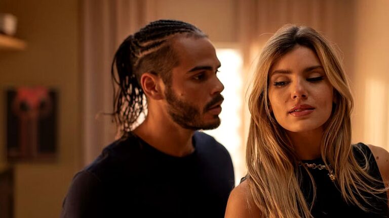 Homem com tranças observa mulher loira com expressão séria em cena dramática ambientada em interior com iluminação quente