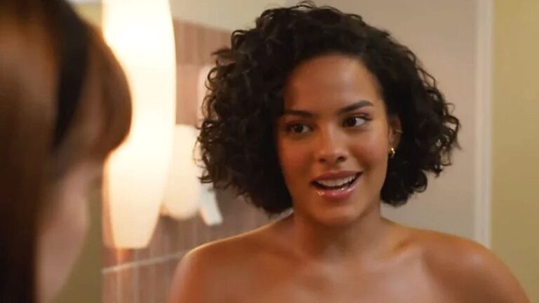 Mulher de cabelo cacheado sorri e conversa com outra pessoa em ambiente interno iluminado