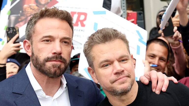 Dois homens posam juntos diante de fãs durante evento público com cartazes e celulares ao fundo