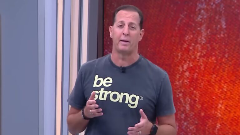 Benjamin Back, apresentador da CNN Brasil, usa camiseta com frase be strong e fala diante de telão no estúdio.