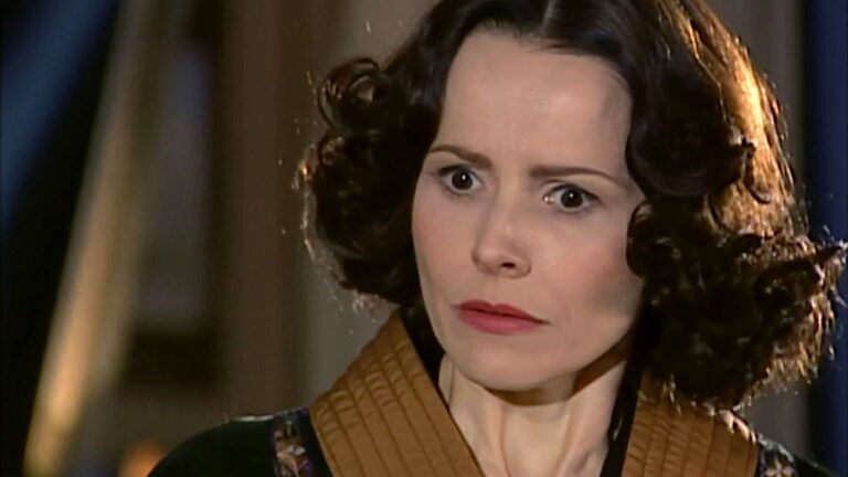 Uma mulher aparece em close, com expressão intensa e desconfiada, sobrancelhas levemente franzidas e olhar fixo em alguém fora de cena. Ela tem cabelos curtos e cacheados, usa maquiagem discreta e veste uma roupa em tons escuros com detalhe em marrom no colarinho. O fundo é desfocado e com iluminação quente, sugerindo uma cena dramática em ambiente interno.