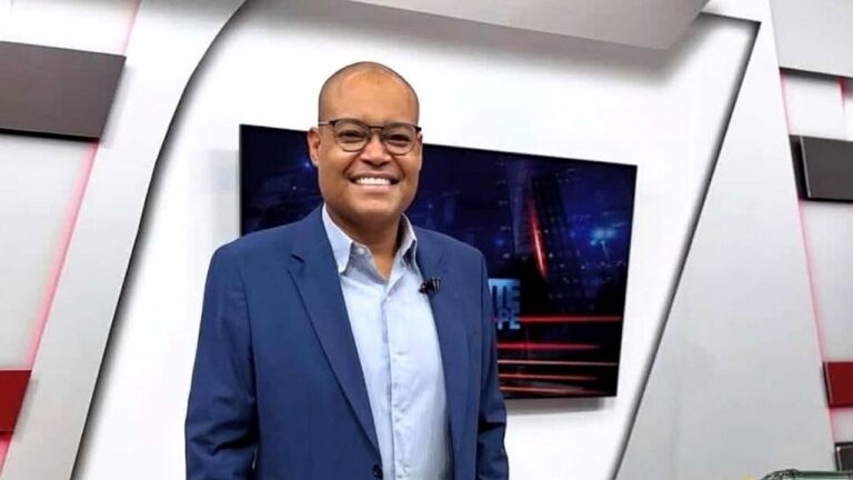 Apresentador sorrindo de terno azul em estúdio de telejornal com telão ao fundo exibindo cenário urbano noturno