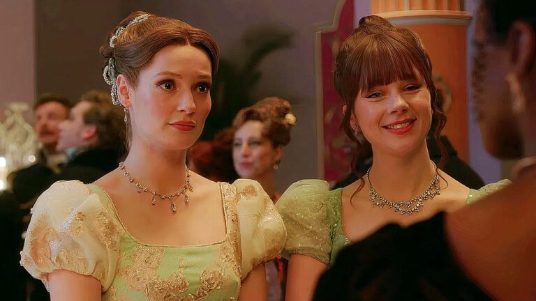 Duas mulheres com vestidos elegantes de época conversam durante baile em salão luxuoso com convidados ao fundo