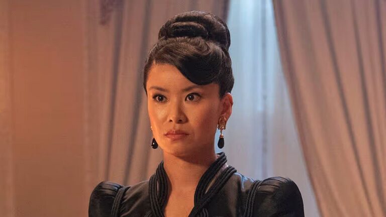 Mulher com penteado elegante e vestido preto observa à frente com expressão séria em ambiente sofisticado com cortinas ao fundo, sugerindo cena de drama ou época