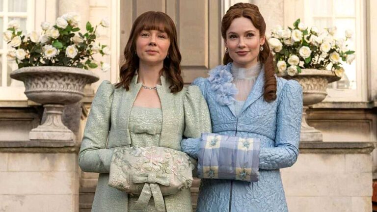 Duas mulheres com vestidos de época posam diante de mansão com arranjos florais em cena de série de época