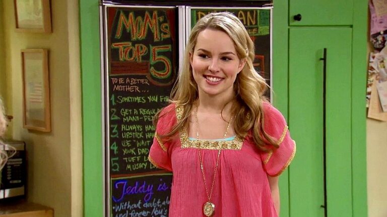 Mulher sorri em cozinha colorida com quadro ao fundo exibindo lista Mom’s Top 5 em cena de série de comédia