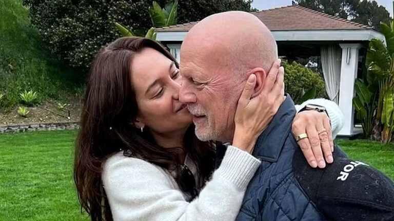 Um homem e uma mulher estão abraçados em um jardim. A mulher segura o rosto do homem enquanto o beija no rosto. Ao fundo há gramado, árvores e uma estrutura semelhante a um gazebo ou área externa de uma casa. A cena transmite um momento de carinho entre o casal.