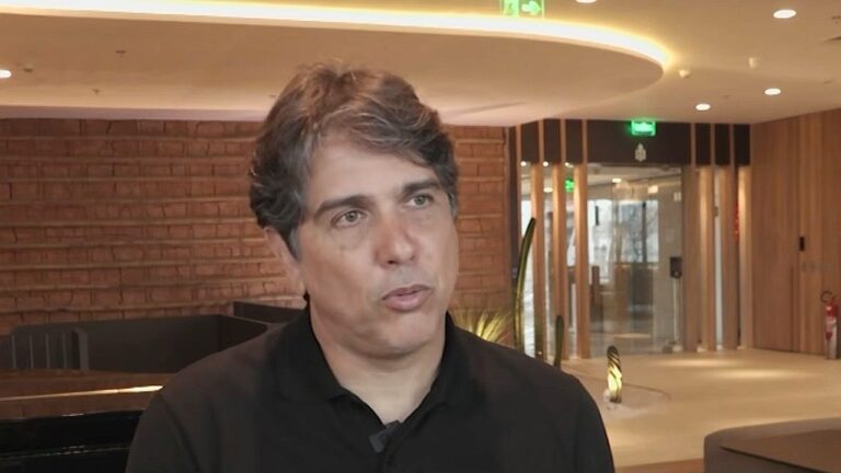 Um homem aparece em um ambiente interno moderno, durante uma entrevista. Ele veste uma camisa escura e tem cabelo curto com fios grisalhos. Sua expressão é séria e concentrada, olhando levemente para o lado enquanto fala. Ao fundo, há um espaço bem iluminado com paredes de tijolos aparentes, iluminação embutida no teto e uma porta de vidro, sugerindo um ambiente corporativo ou hotel.