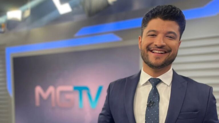 Jornalista Cadu Alvim sorridente, veste terno azul escuro e gravata de poá, posando em estúdio de TV com o logotipo do MGTV ao fundo da imagem