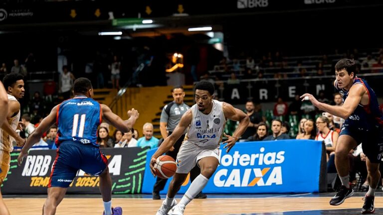 Uma partida de basquete está em andamento dentro de um ginásio. No centro da imagem, um jogador com uniforme claro dribla a bola enquanto avança, sendo marcado por adversários com uniformes azuis. Outros jogadores se posicionam ao redor, atentos à jogada. Ao fundo, há arquibancadas com público e placas de publicidade, indicando um jogo profissional ou competitivo.