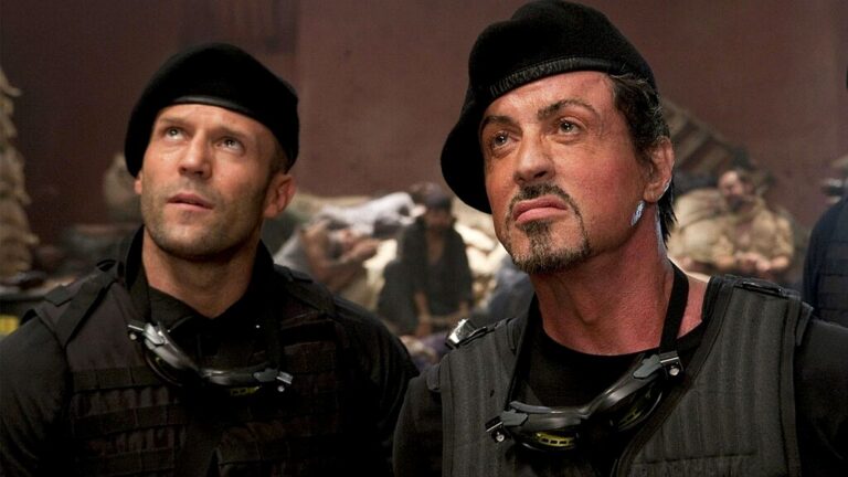 Dois homens usando boinas pretas e coletes táticos observam algo acima com expressão séria em cenário de ação com destroços ao fundo