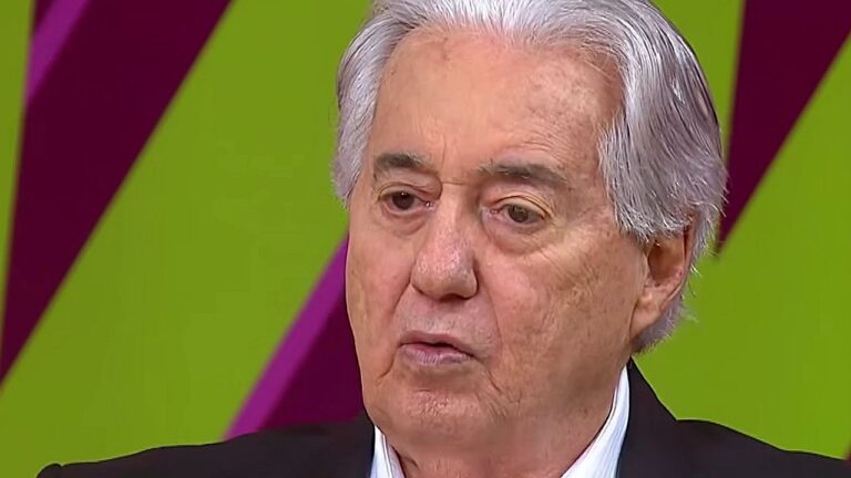 Homem idoso de cabelos brancos aparece em close durante participação em programa de televisão, com expressão séria enquanto fala; ao fundo, um cenário colorido de estúdio.