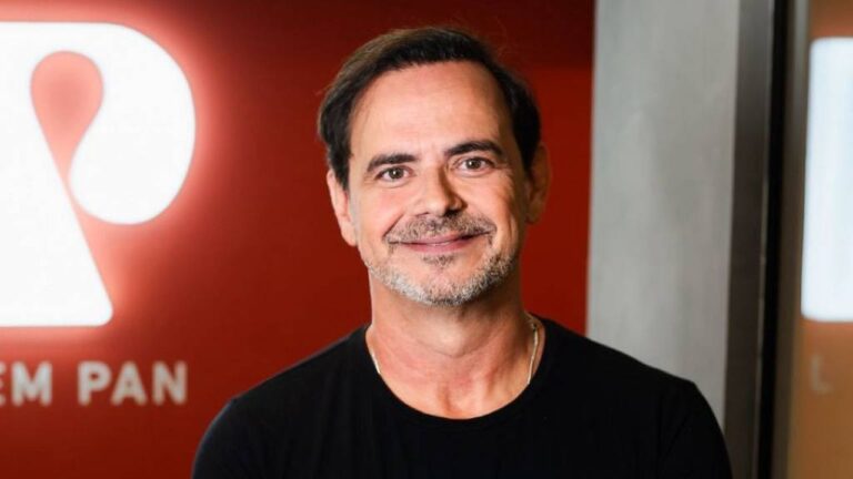 Márvio Lúcio, o Carioca, sorri para a foto usando camiseta preta, posicionado à frente do logotipo iluminado da Jovem Pan em uma parede vermelha