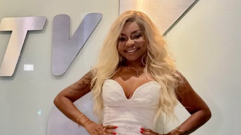 Cariúcha de vestido branco tomara que caia e longos cabelos loiros sorri com as mãos na cintura em frente a uma parede branca com o logotipo da RedeTV!