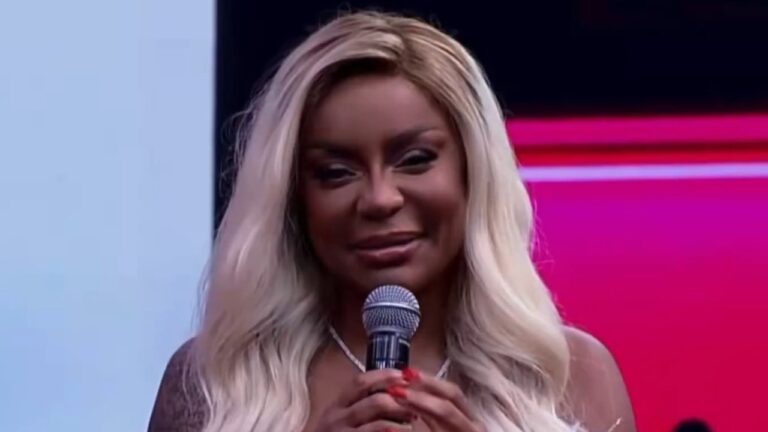 Cariúcha com cabelo loiro longo segura microfone e fala no palco do Superpop em estúdio com fundo iluminado em rosa