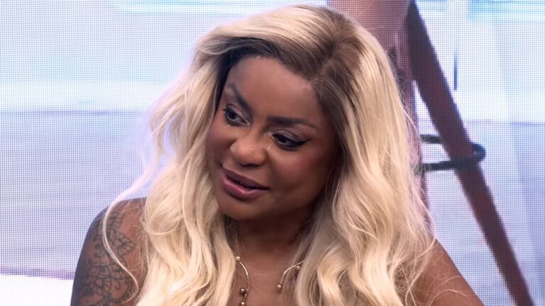 Apresentadora Cariúcha com cabelos loiros longos e ondulados, usa colar dourado e exibe expressão de emoção com lágrimas nos olhos em cenário de estúdio