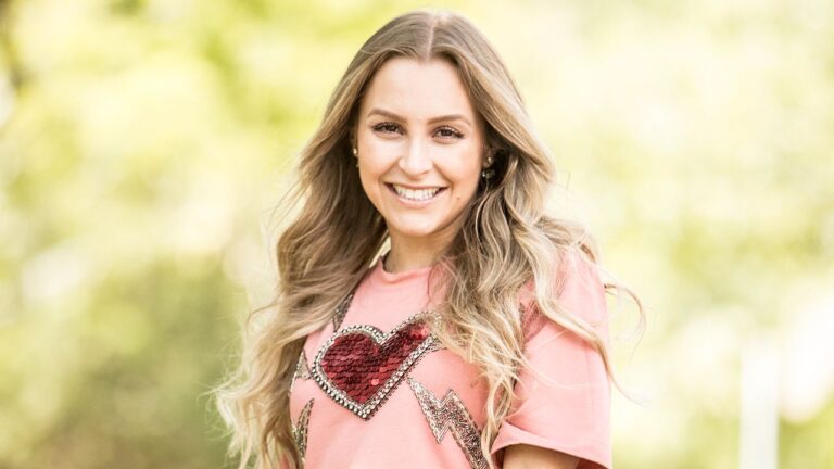 Atriz Carla Diaz sorri com cabelos loiros ondulados, vestindo camiseta rosa com coração de lantejoulas vermelhas, em plano médio com fundo verde desfocado