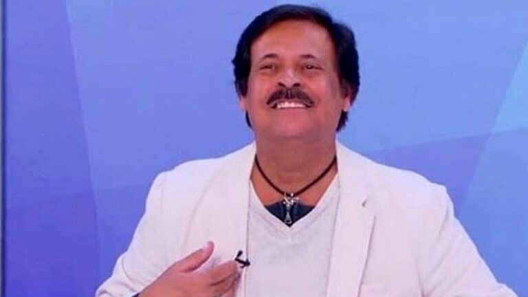 Homem de bigode veste blazer branco e sorri enquanto fala em estúdio de televisão com fundo azul iluminado.