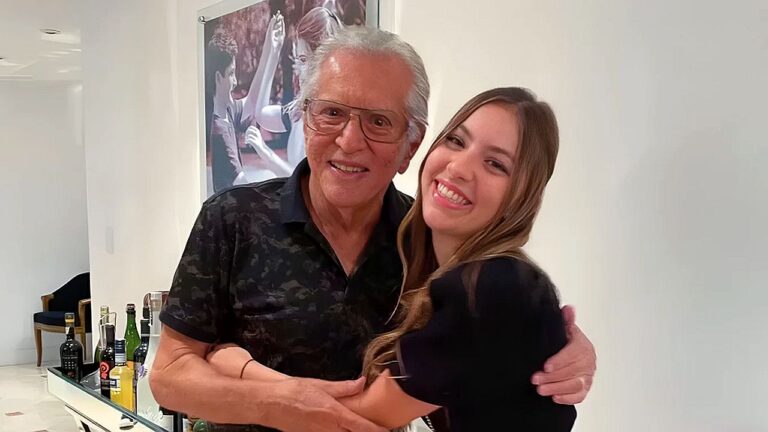 Texto Alternativo disse: Homem idoso de cabelos brancos e óculos abraça uma jovem sorridente em ambiente interno, posando para foto diante de um aparador com garrafas e um quadro decorativo na parede.