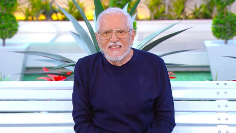 Homem idoso de barba branca e óculos fala diante das câmeras em cenário de programa de televisão com plantas decorativas ao fundo