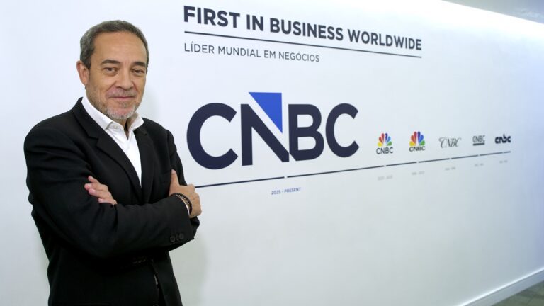 Carlos José Marques, de terno preto, posa com os braços cruzados em frente a uma parede branca com o logotipo do Times Brasil | CNBC. Ao fundo, lê-se “First in Business Worldwide” e “Líder mundial em negócios”, além de diferentes versões do logotipo da emissora exibidas em linha.