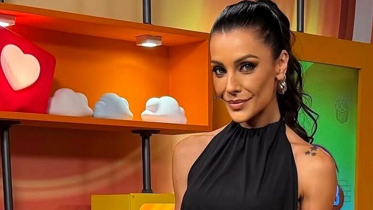 Mulher com cabelo escuro preso em rabo de cavalo posa em estúdio colorido, usando vestido preto sem mangas e brincos; ao fundo há prateleiras decorativas com objetos em formato de coração e nuvens.