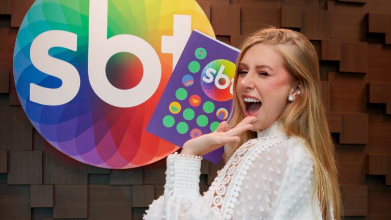 Influenciadora Cela Lopes sorri e segura envelope roxo ao lado do logotipo colorido do SBT, vestindo blusa branca rendada em frente a parede de madeira